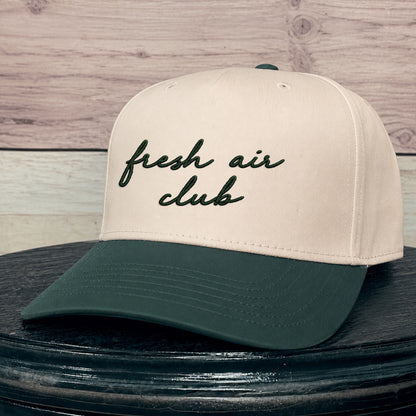 A baseball cap with an natural crown and a dark green brim, featuring the text 'fresh air club' embroidered in dark green script on the front. The cap is displayed on a dark wooden surface against a light wood plank background.
