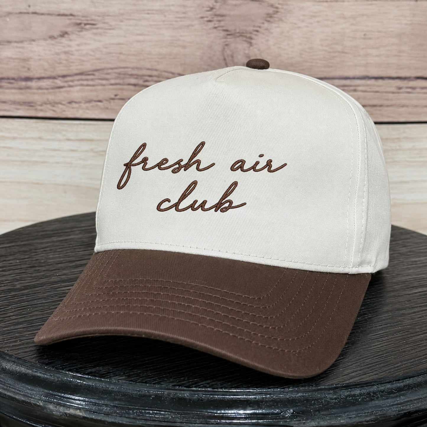 A baseball cap with an off-white crown and a brown curved brim, featuring the words 'fresh air club' embroidered in brown script on the front. The cap is displayed on a dark round surface with a light wood plank background.