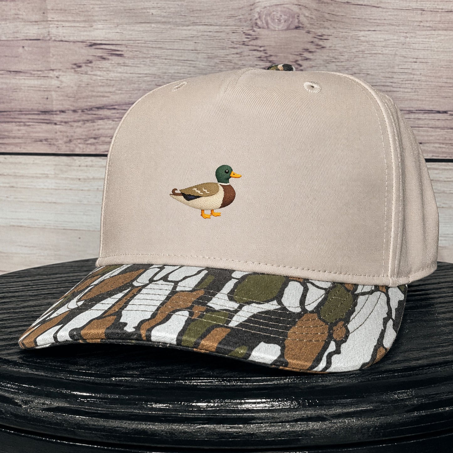 A beige baseball cap with a small embroidered mallard duck on the front and a camouflage tree bark patterned visor, displayed on a black surface against a rustic wood plank background.