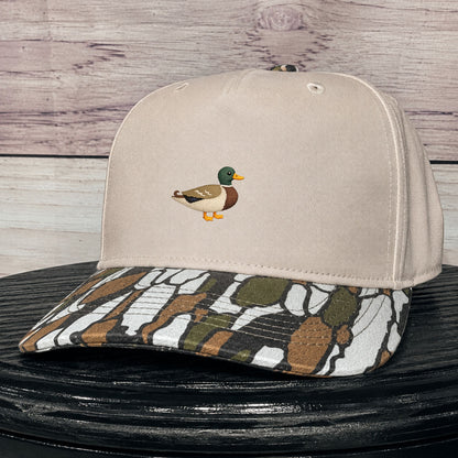 A beige baseball cap with a small embroidered mallard duck on the front and a camouflage tree bark patterned visor, displayed on a black surface against a rustic wood plank background.