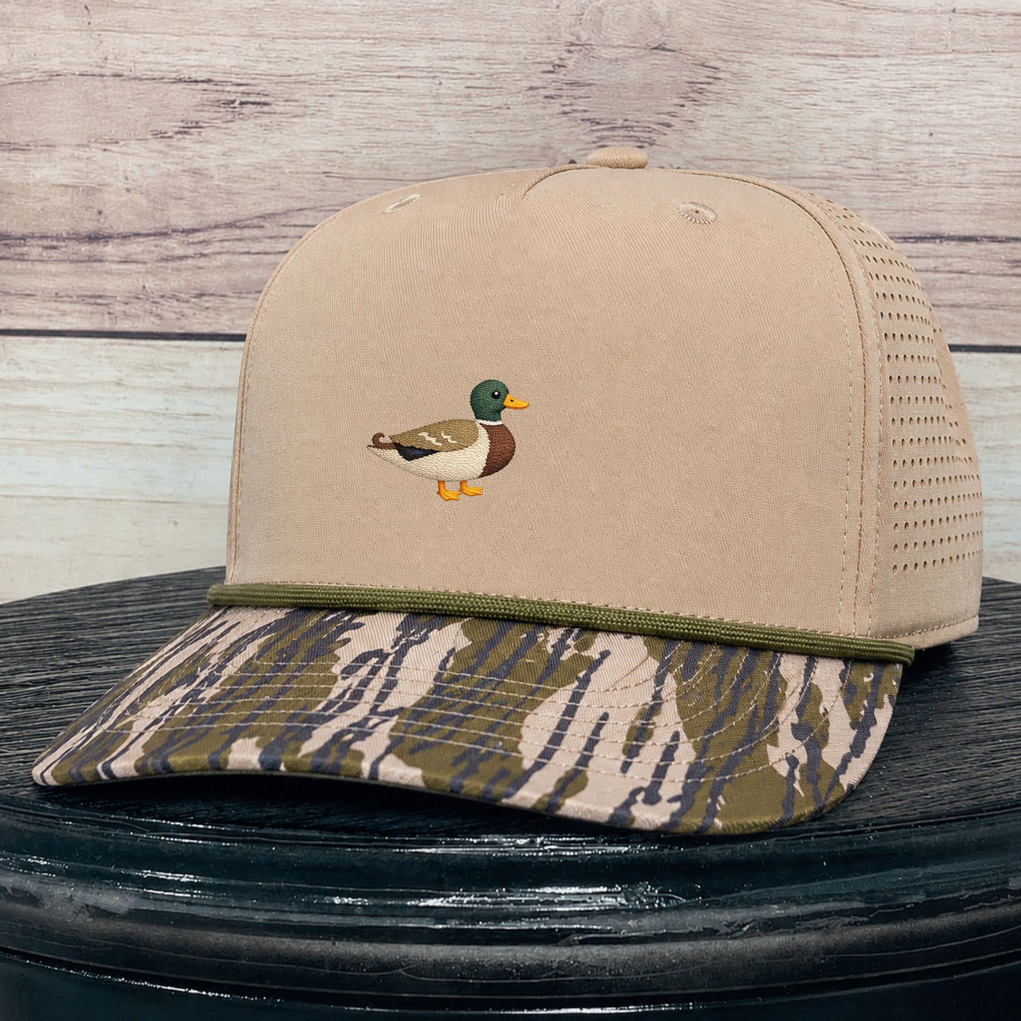 A beige trucker-style baseball cap with a small embroidered mallard duck on the front, a green trim line, and a camouflage-patterned brim, displayed on a dark round surface against a light wood plank background.