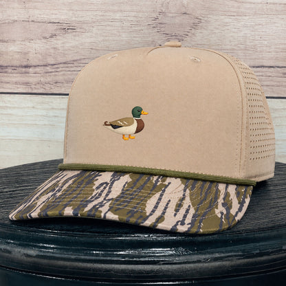 A beige trucker-style baseball cap with a small embroidered mallard duck on the front, a green trim line, and a camouflage-patterned brim, displayed on a dark round surface against a light wood plank background.