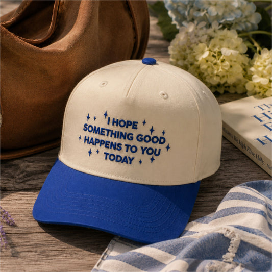 A cream and royal blue baseball cap with the embroidered text 'I HOPE SOMETHING GOOD HAPPENS TO YOU TODAY' and small star accents, styled on a wooden surface with a brown bag, flowers, and a striped cloth nearby.